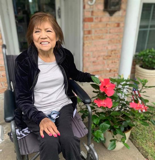 A smiling elderly woman in a wheelchair enjoys the sun next to colorful garden flowers.