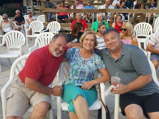 Three friends share laughter and drinks while seated at a casual beachside event during sunset.