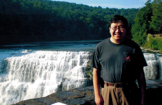 A person stands happily beside a waterfall, surrounded by lush greenery and clear blue skies.
