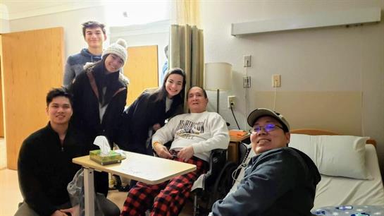 Family smiles as they gather around a man in a wheelchair in a hospital room.