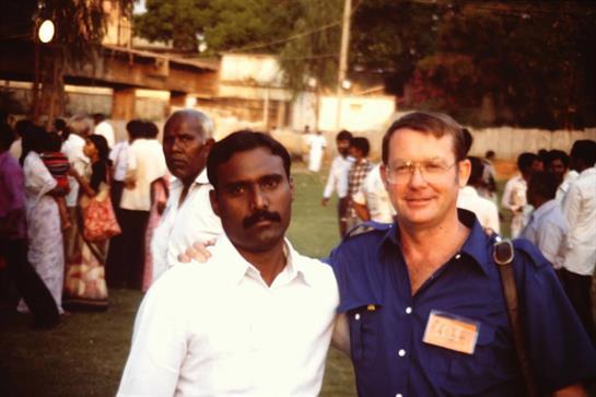 Two men stand side by side, smiling, among a crowd at a social event in India during late afternoon.