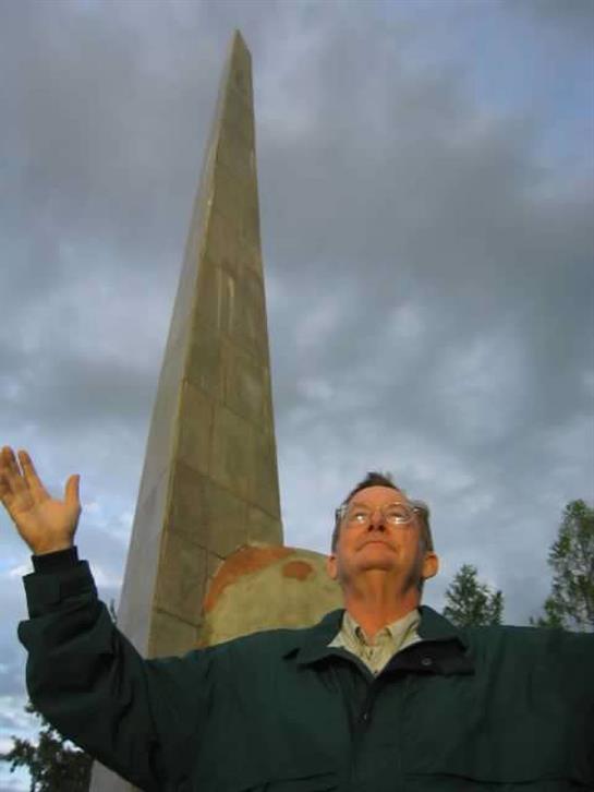 An elderly man stands with arms outstretched, facing a towering obelisk amidst overcast skies.