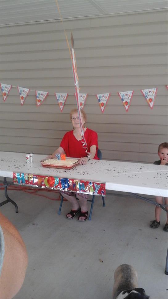 An elderly woman in a red shirt sits at a party table with a child, celebrating.