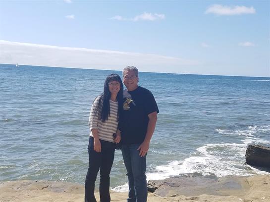 Two people stand close together on a sandy beach, smiling and enjoying a sunny day by the ocean.