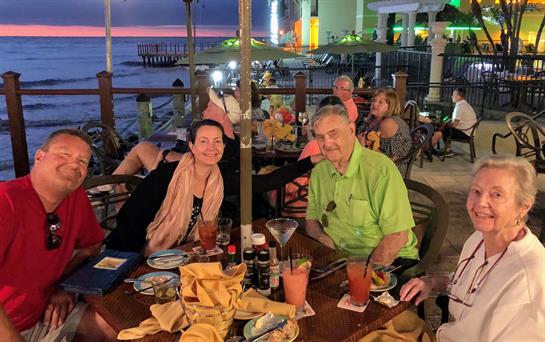 Group of friends shares laughter and drinks at a vibrant seaside restaurant during sunset.