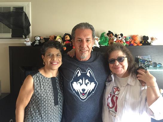 Three friends stand together in a bright room with various plush toys, smiling happily.