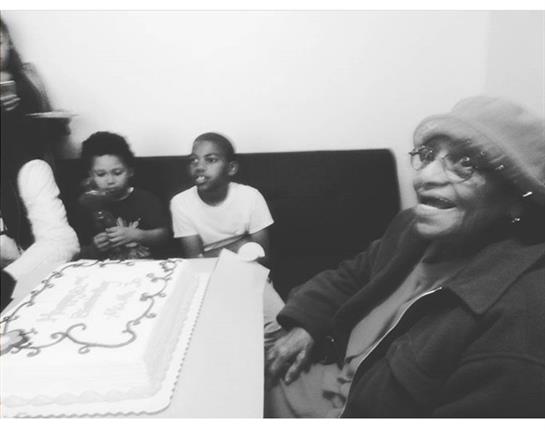 An elderly man smiles at a birthday party with children gathered around a decorated cake.