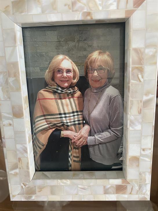Two women smile while holding hands, dressed warmly, in a friendly indoor atmosphere.