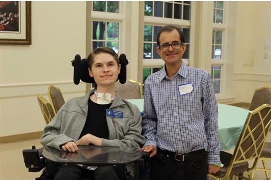 A young man with a communication device smiles alongside an adult mentor during a gathering.