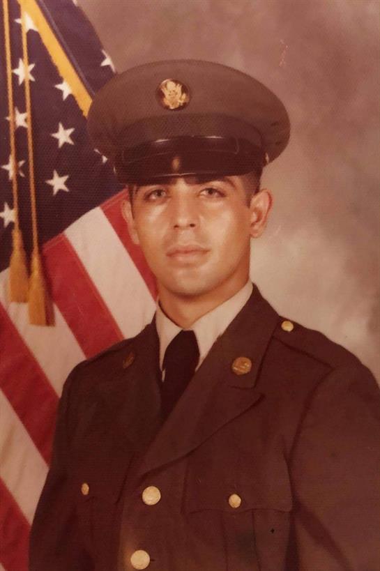 A young soldier stands in uniform, displaying pride with the American flag behind him.