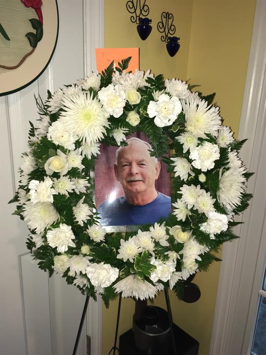 White flowers frame a smiling man's portrait, honoring his memory with a heartfelt tribute.
