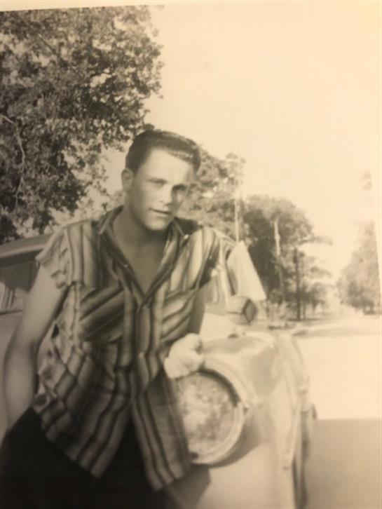 A young man poses with a classic car surrounded by trees, enjoying a sunny afternoon.