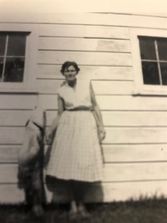 A woman in a light-colored dress poses with a young child beside a wooden house.