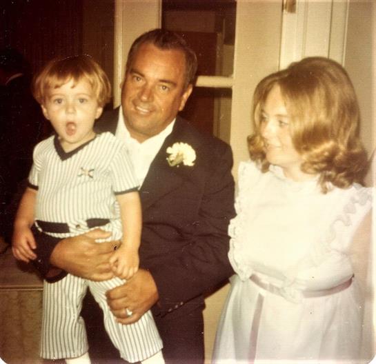 A man holds a child in stripes, while a woman in a light dress stands beside them.