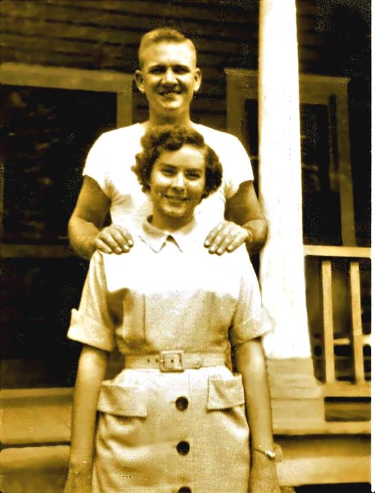 A man and woman stand together outdoors, smiling warmly in front of a house from the mid-1900s.