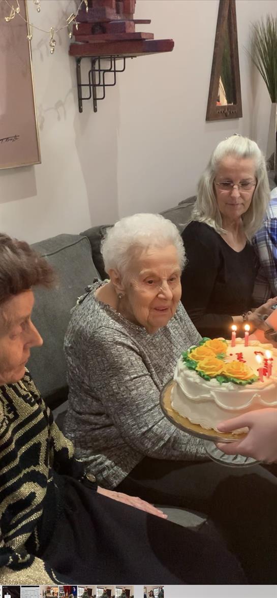 Family members gather in a living room to celebrate a centenarian's birthday with cake and candles.