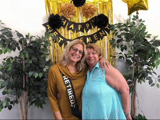 Two women pose joyfully during a retirement celebration surrounded by decorations.