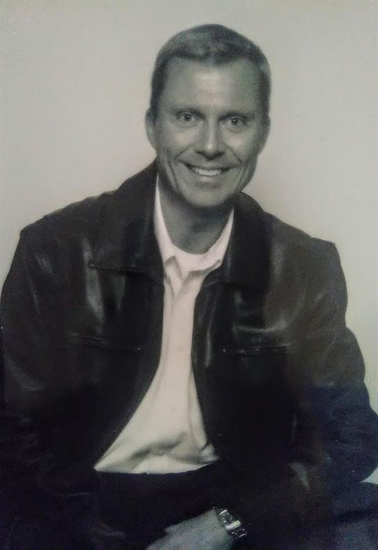 A man with a friendly smile sits wearing a leather jacket and white shirt in a plain background.