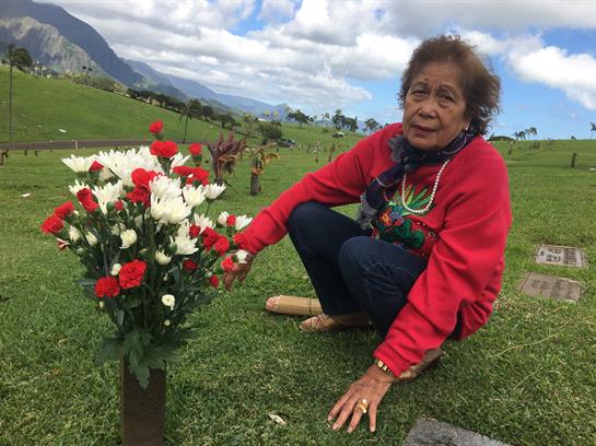 An elderly woman arranges flowers at a gravesite, with mountains and clear skies around her.