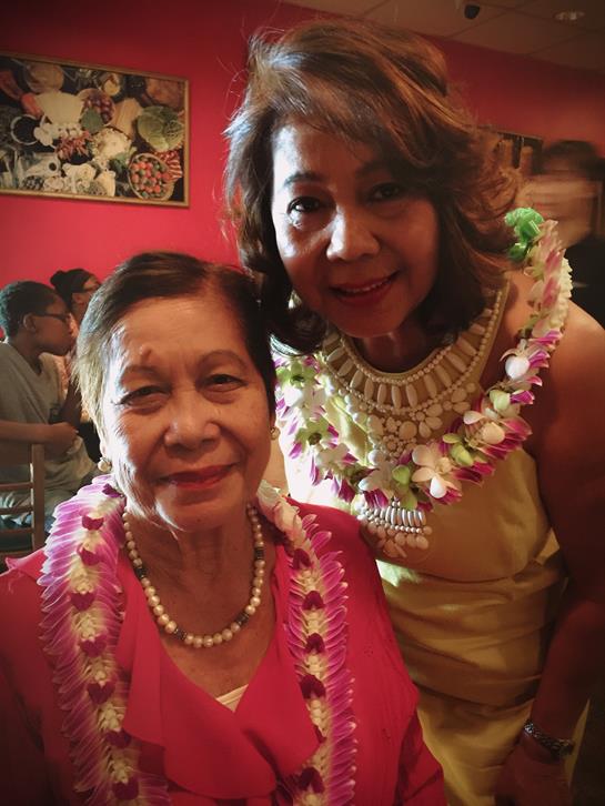 Two women smile warmly while adorned with colorful leis in a festive restaurant atmosphere.