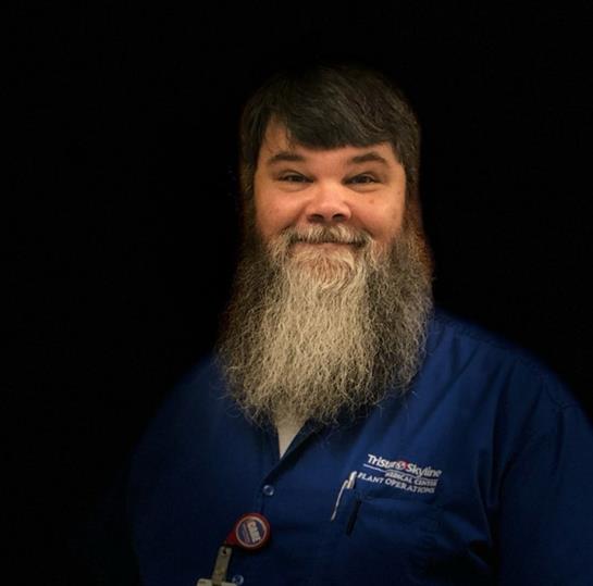 A friendly male healthcare worker with a long beard smiles warmly in a dark setting.