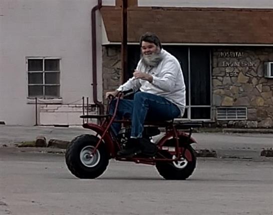 A person rides a small motorcycle around an urban setting, smiling and waving at onlookers.