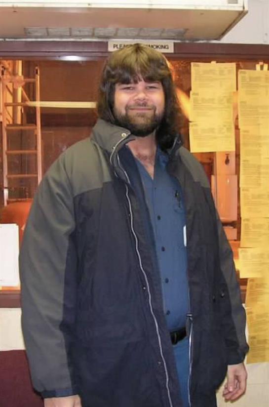 Man wearing a warm jacket smiles while standing indoors near cluttered notes and papers.