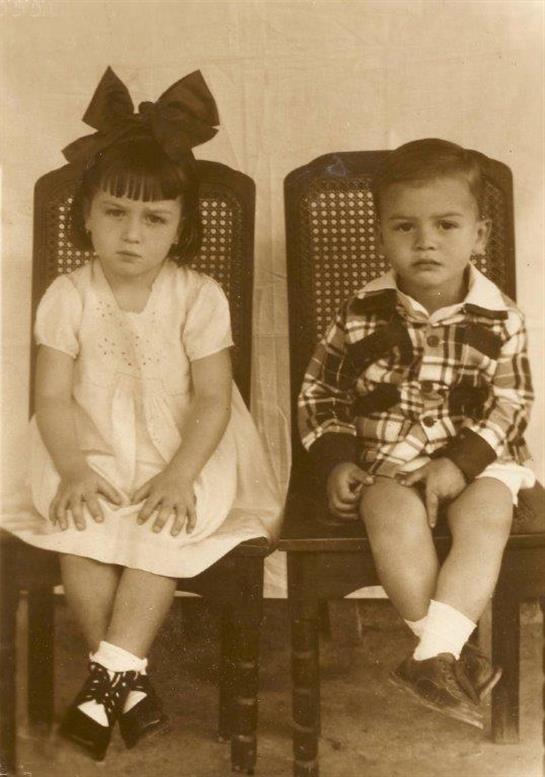 Two young children sit quietly on chairs, wearing classic outfits with an old-fashioned backdrop.