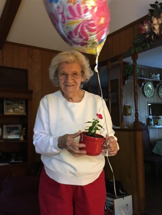 An elderly woman smiles while holding a red flower pot and a colorful balloon indoors.