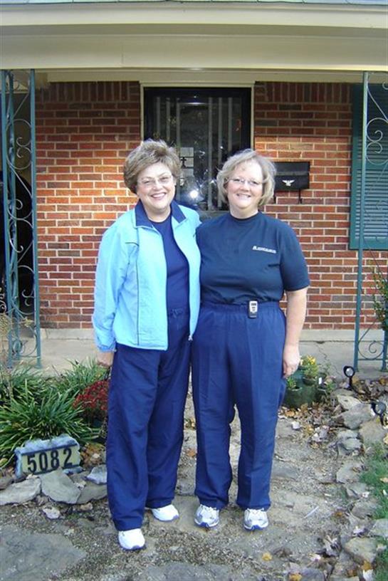 Two women stand close together, smiling in casual blue outfits in front of a brick home.
