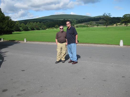 Two friends stand together on a concrete path surrounded by a lush green landscape.