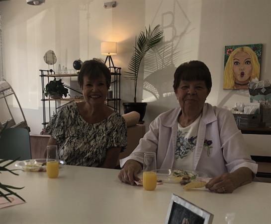 Two women sit at a table, smiling while enjoying drinks and snacks in a well-lit, stylish room.