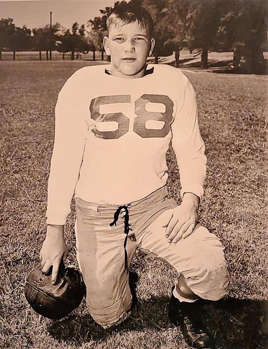 A young football player kneels on a grassy field wearing a classic uniform while holding a ball.