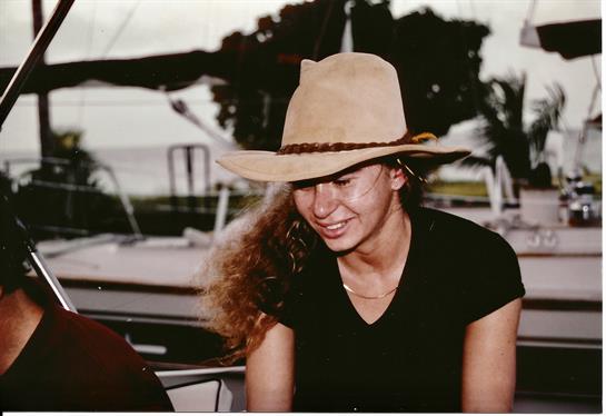 A cheerful woman in a hat enjoys a sunny day at the marina with nearby boats.