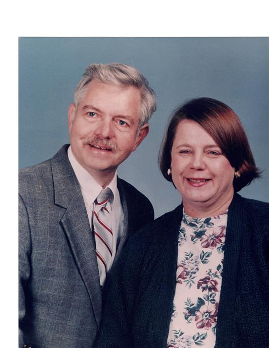 Two individuals stand side by side, smiling warmly in a formal attire against a neutral backdrop.