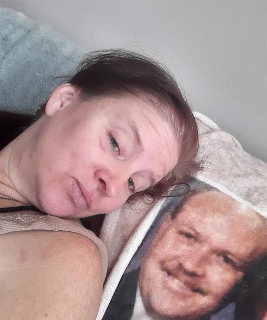 A woman is playfully posing beside a smiling man's portrait on a cushion at home.