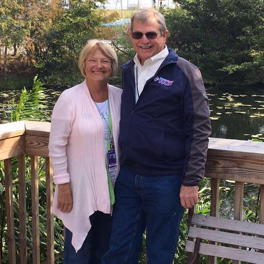 A cheerful couple stands close together smiling beside a pond surrounded by lush plants.