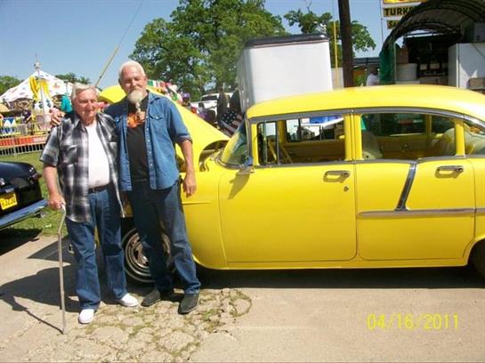 Two men stand proudly beside a vintage yellow car at a local automotive gathering on a sunny day.