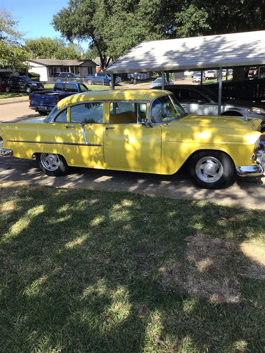 A bright yellow vintage car rests in a shady spot while others line the street nearby.