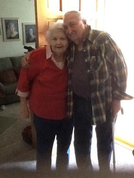 An elderly couple smiles warmly while standing together in a cozy living room setting.