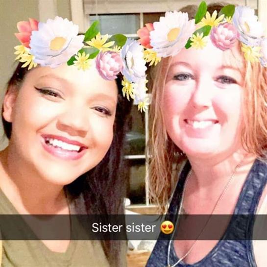 Two women smile brightly for a fun selfie wearing floral crowns in a cozy setting.
