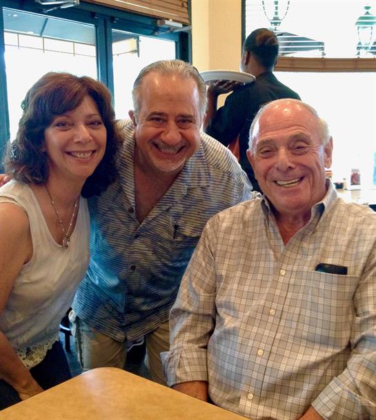 Three friends smile together while enjoying food and conversation in a restaurant setting.