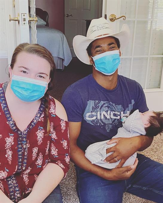 Two individuals smile while holding a baby, all wearing protective masks in a home environment.