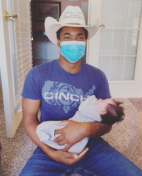 A man with a cowboy hat and mask cradles an infant in a cozy indoor setting.