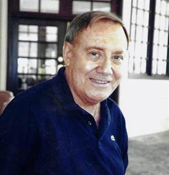 Elderly man with a warm smile sits comfortably indoors near large windows during daylight.