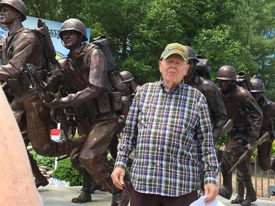 Elderly man in plaid shirt stands near a bronze soldier statue, reflecting on history and service.