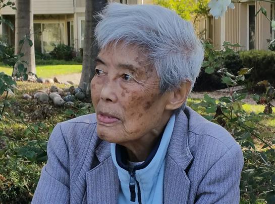 An elderly individual with gray hair relaxes in a garden, observing the scenery under the sun.