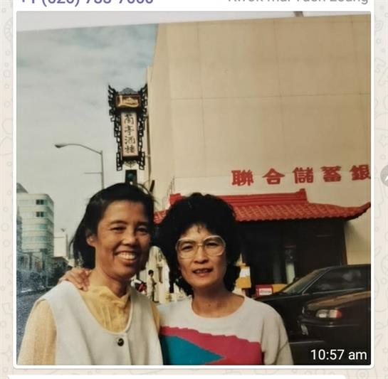 Two women smile together on a city street, surrounded by buildings and parked cars in the daytime.