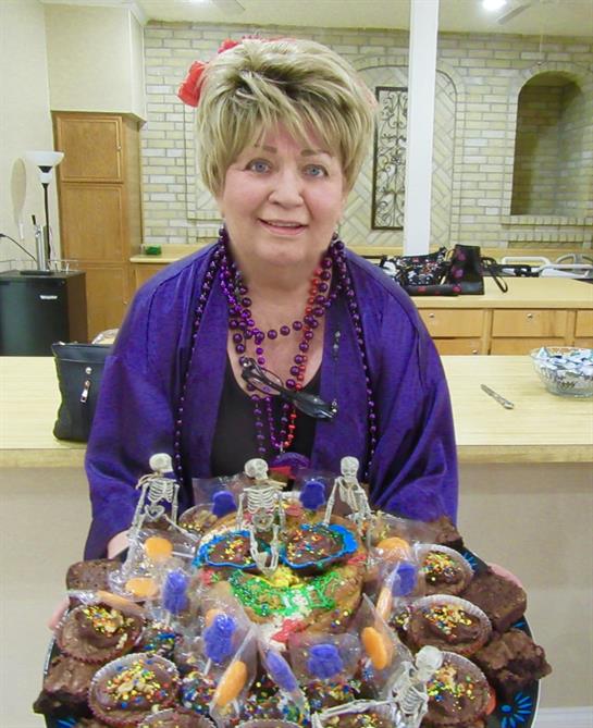 A cheerful woman holds a vibrant tray of decorative bottles and treats in her kitchen.