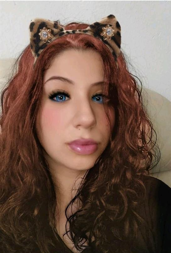 A young woman with wavy brown hair and blue eyes looks relaxed at the camera indoors.